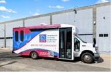 A mobile blood donation center vehicle is parked outside a building under a blue sky with scattered clouds.