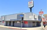 Gray corner building with "Optique Eye" signage, empty sign frames, and red "For Lease" banners, located at a street intersection on a clear day.