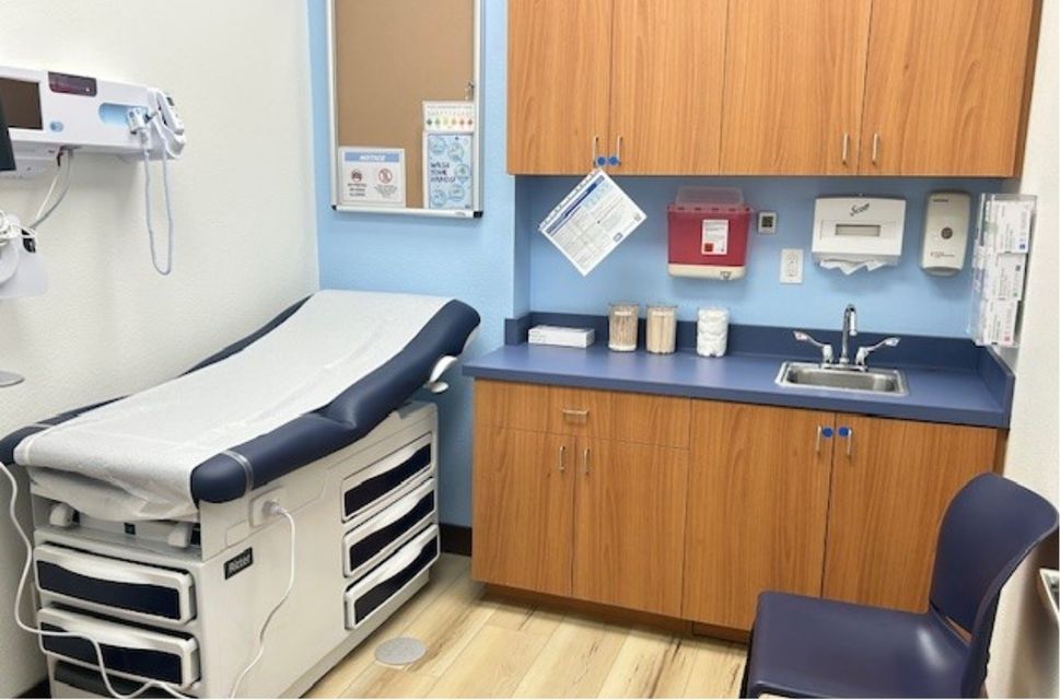 A clean medical exam room with an exam table, wooden cabinets, countertop with sink, supplies, and medical equipment on the wall.